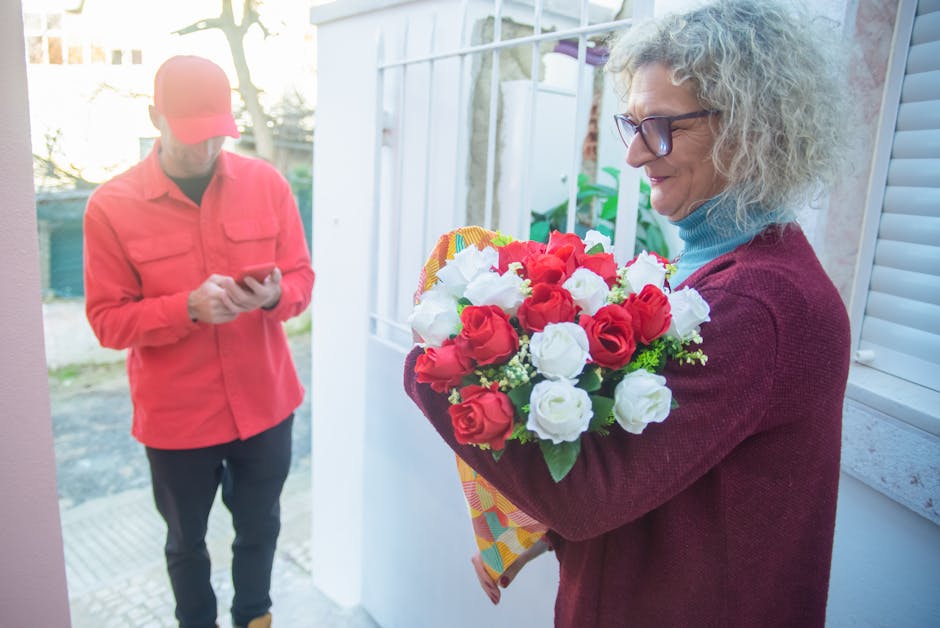 An elderly woman with curly gray hair and glasses, dressed in a maroon cardigan and a blue turtleneck, is holding a vibrant floral bouquet composed of red and white roses, accented with green foliage,
