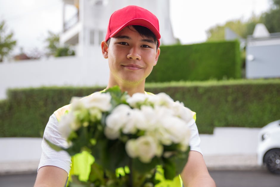 A young delivery person wearing a red cap and a high-visibility vest, smiling proudly as they extend a bouquet of fresh white roses and greenery towards the camera, with a blurred residential street a