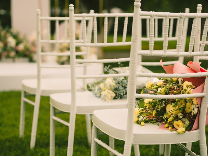 A row of white wooden chairs arranged outdoors on a green lawn, with two floral arrangements placed on the seats. The arrangements feature yellow roses, white baby's breath, and greenery, wrapped in p