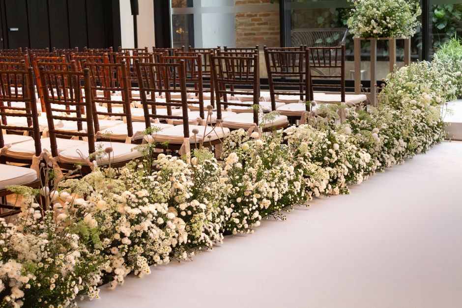 A spacious indoor wedding ceremony area decorated with a long, lush floral arrangement of white baby's breath and small white roses running along the front edge of the aisle. Behind the flowers, there
