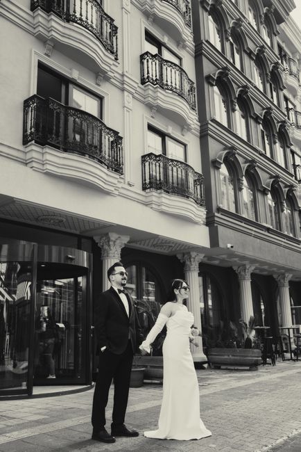 A black-and-white photograph depicting a bride and groom standing hand in hand outdoors in front of an ornate, multi-story building with decorative iron balconies and large windows. The bride wears a