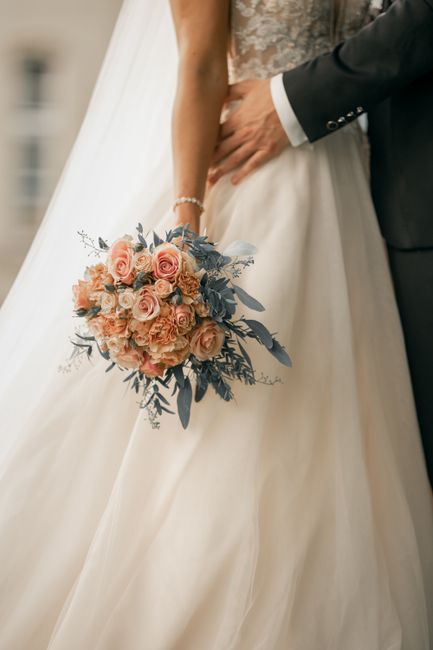 A bride and groom standing close together, with the bride holding a large, round bouquet of pastel pink roses, peachy-orange roses, and dusty blue-green foliage, arranged in a lush, cascading style. T