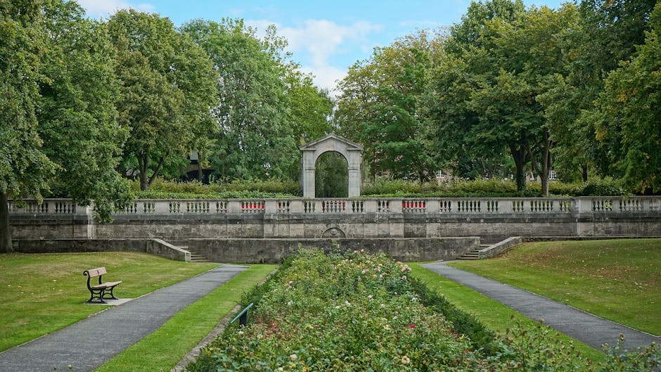 A scenic park scene featuring a symmetrical flower bed filled with a variety of blooming flowers, including roses and daisies, flanked by two paved walkways. On the left, there is an empty wooden park