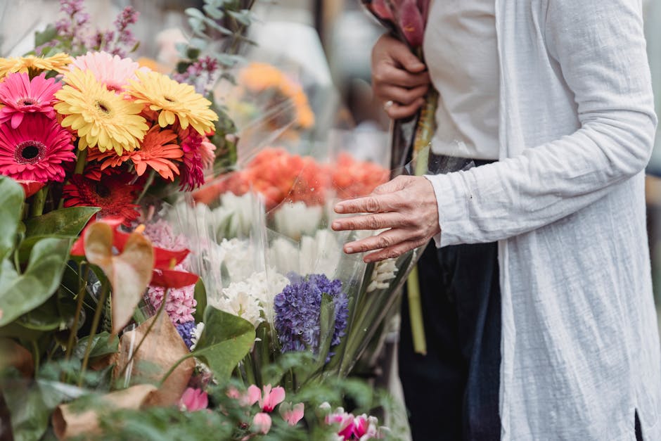 A person in a light-colored coat is selecting fresh flowers from a vibrant floral display at a florist. The arrangement includes colorful gerbera daisies in pink, yellow, and orange hues, alongside pu